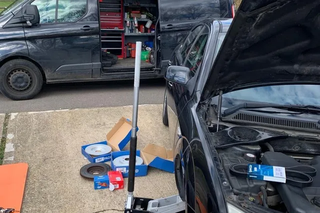 A van with its side doors open showing tool drawers, parked beside a car with its bonnet raised, a trolley jack and brake discs on the ground